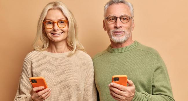 horizontal-shot-of-elderly-couple-use-modern-technologies-hold-smart-phones-reads-text-messages-connected-to-wireless-internet-wear-casual-jumpers-isolated-over-brown-wall